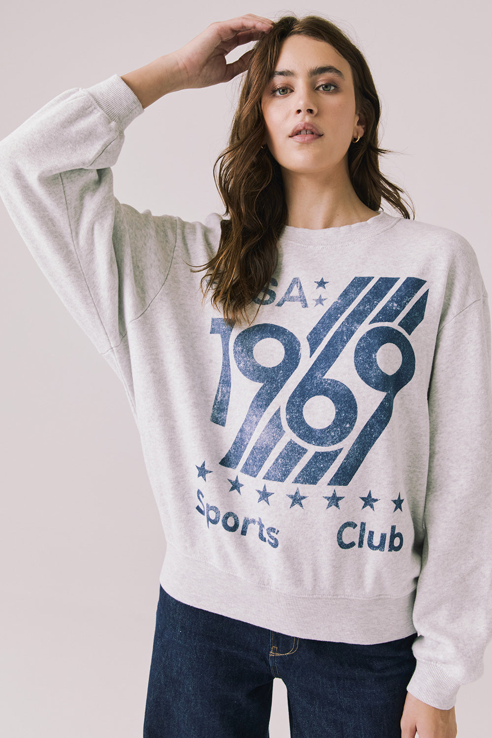 Front view of woman wearing Chaser’s light grey USA Sports Club Long Sleeve with a blue USA 1969 graphic on a white background