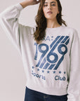 Front view of woman wearing Chaser’s light grey USA Sports Club Long Sleeve with a blue USA 1969 graphic on a white background