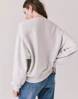 Back view of woman wearing Chaser’s Star Chainstitch Light Grey Long Sleeve on a plain background