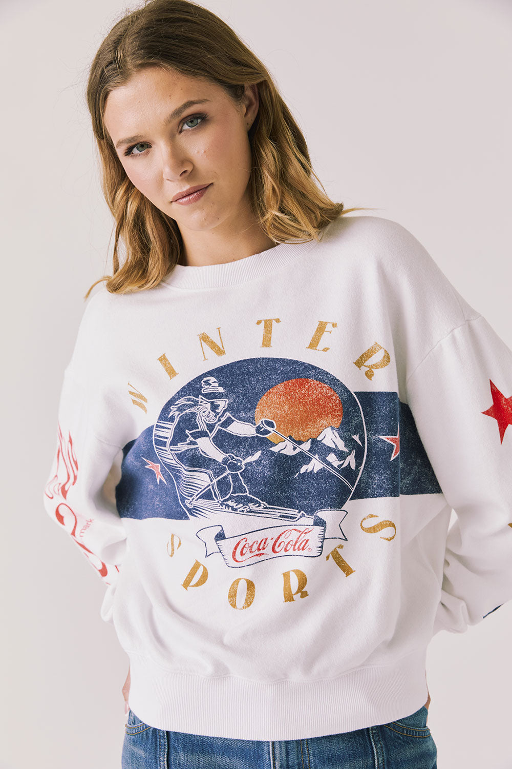 Front view of woman wearing Chaser’s white Coca-Cola Winter Sports Long Sleeve with vintage-inspired graphic design and blue jeans on a plain background