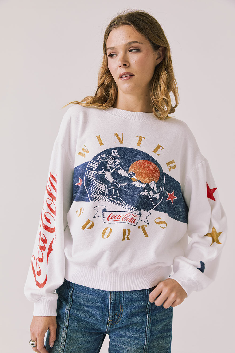 Front view of woman wearing Chaser’s white Coca-Cola Winter Sports Long Sleeve with vintage-inspired graphic design and blue jeans on a plain background