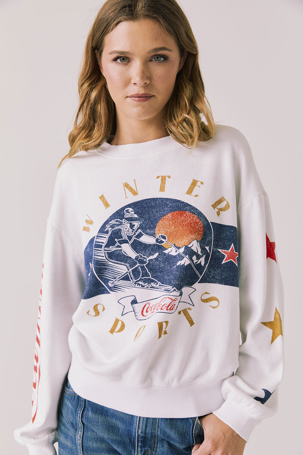 Front view of woman wearing Chaser’s white Coca-Cola Winter Sports Long Sleeve with vintage-inspired graphic design and blue jeans on a plain background