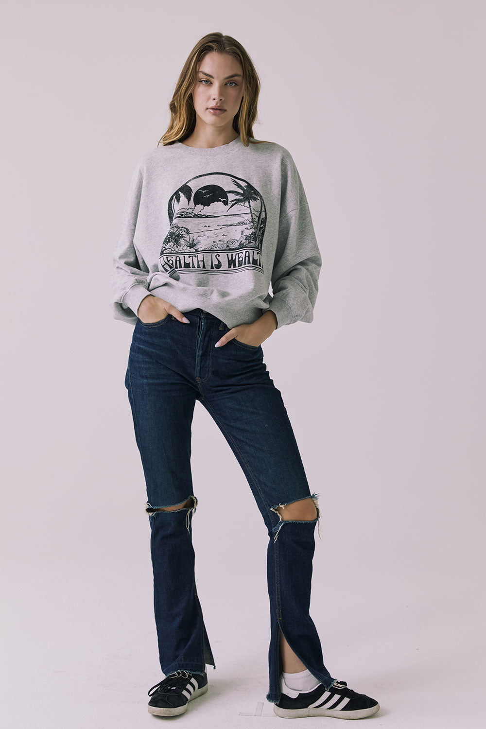 Full body front view of a woman wearing Chaser's gray Health Is Wealth Long Sleeve with a graphic design and ripped denim jeans on a plain background