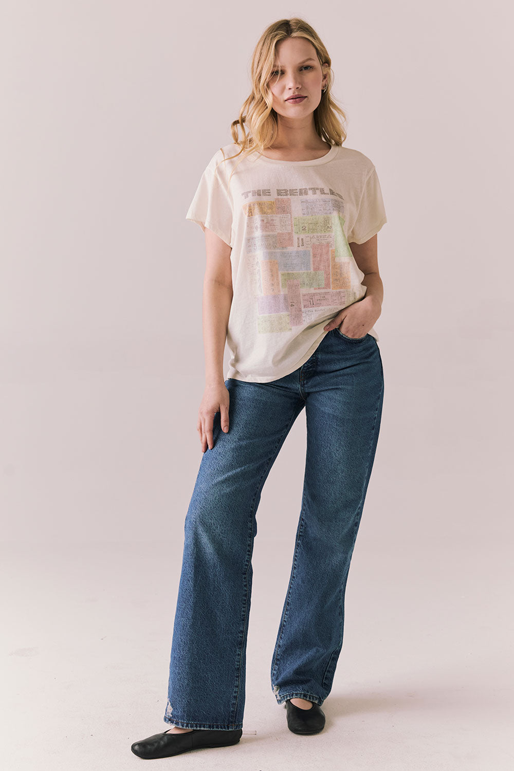 Full body view of model wearing The Beatles Ticket Stub Collage Tee in Starry White with vintage ticket graphic and classic band logo and blue jeans standing  against a plain background