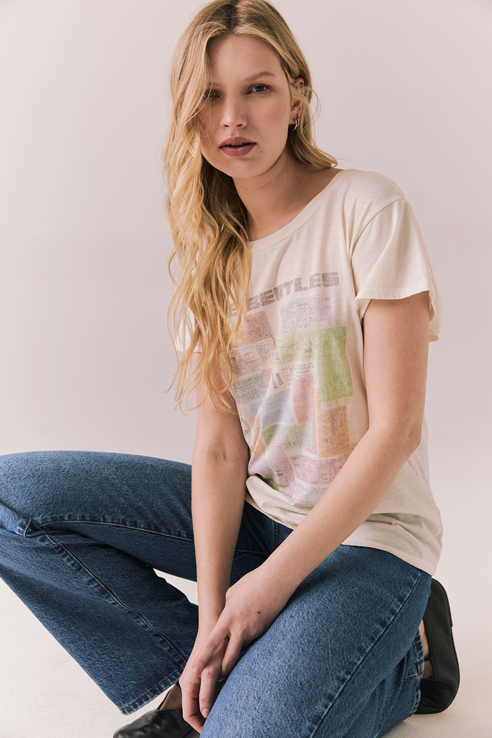 Full body view of model wearing The Beatles Ticket Stub Collage Tee in Starry White with vintage ticket graphic and classic band logo and blue jeans crouched on the floor against a plain background