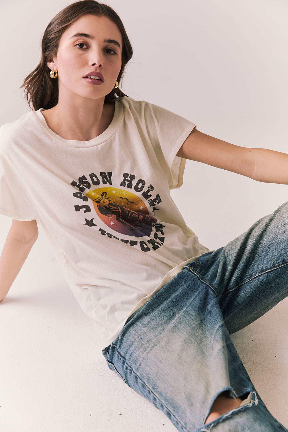 Model wearing the Jackson Hole Sunset Tee in white with scenic Western graphic with graphic design and ripped jeans  sitting on a plain background