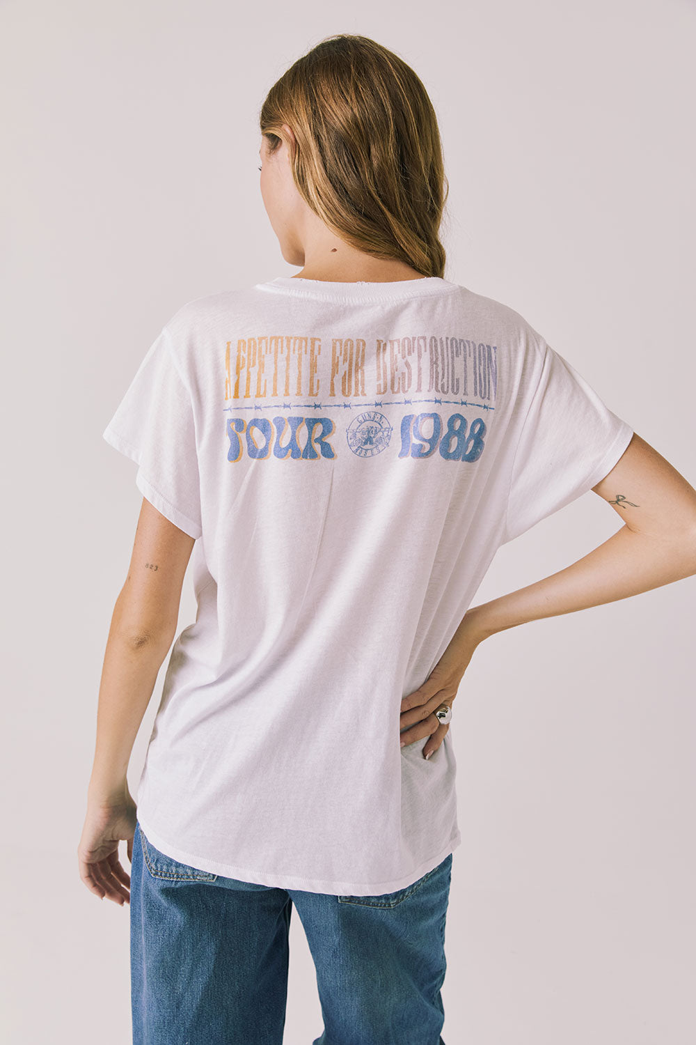 Back view of woman wearing Chaser’s white Guns N’ Roses World Tour Tee with “Appetite for Destruction Tour 1988” text (licensed band collaboration) on a plain background