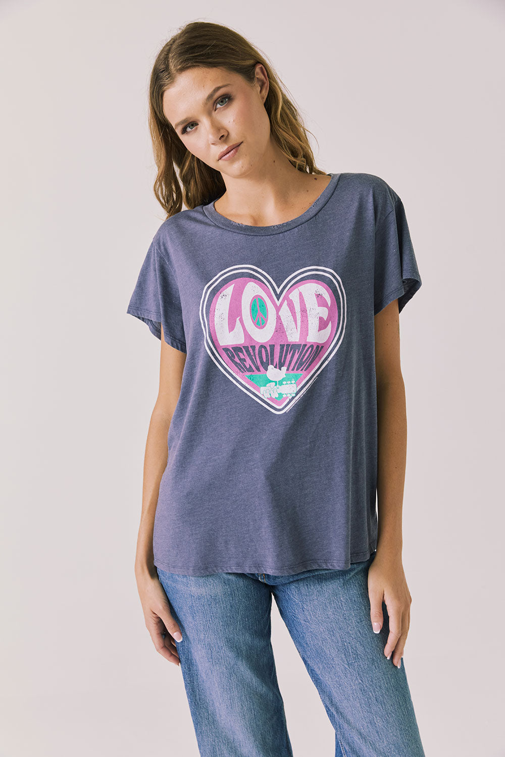 Front view of woman wearing Chaser’s slate blue Woodstock Love Revolution Tee with pink heart design and ‘LOVE’ text on a plain background (licensed music collaboration)