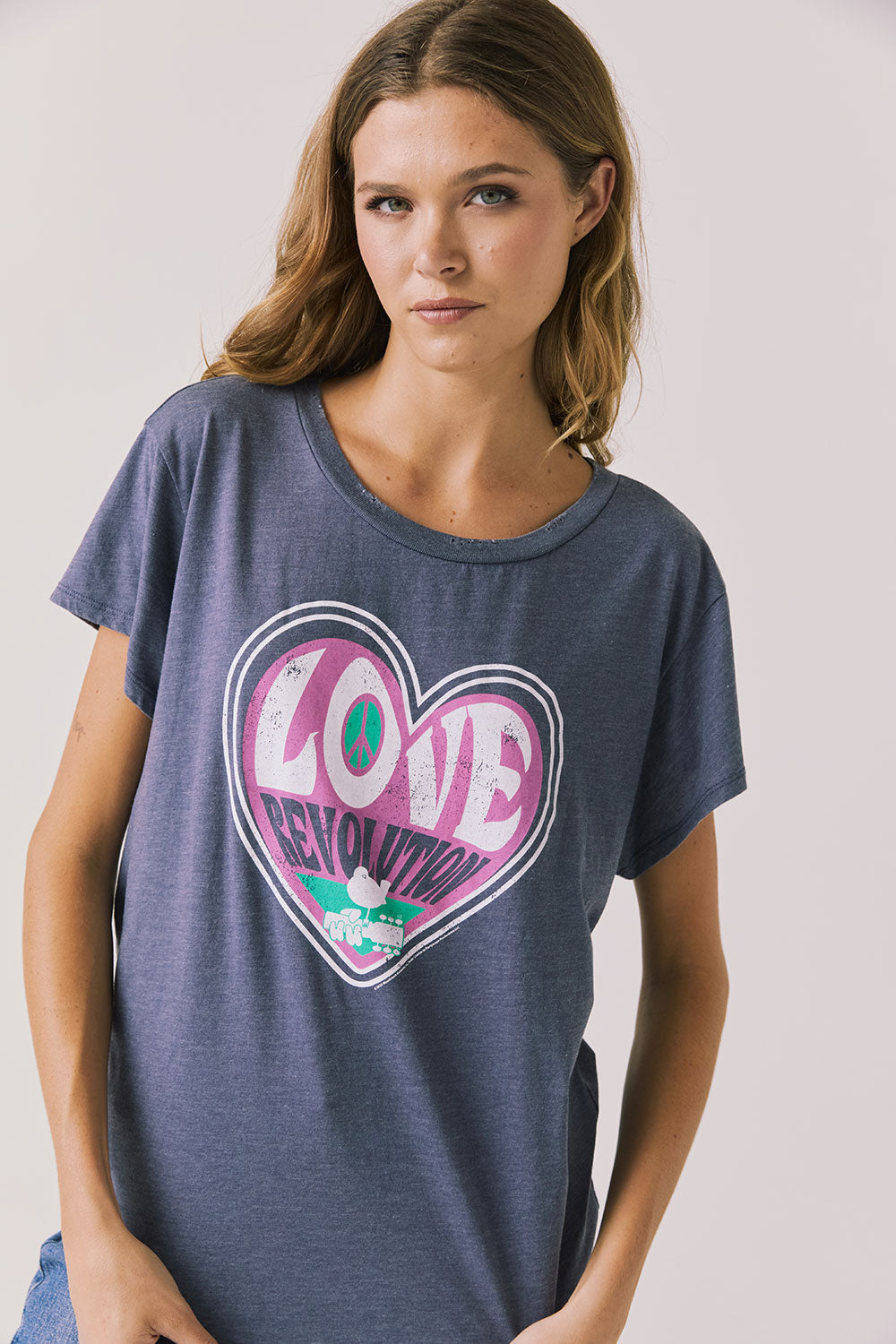 Front view of woman wearing Chaser’s slate blue Woodstock Love Revolution Tee with pink heart design and ‘LOVE’ text on a plain background (licensed music collaboration)