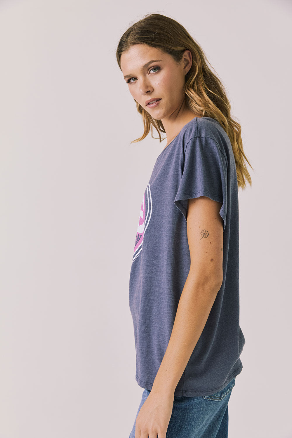 Side view of woman wearing Chaser’s slate blue Woodstock Love Revolution Tee showing pink heart and text detail on a plain background (licensed music collaboration)