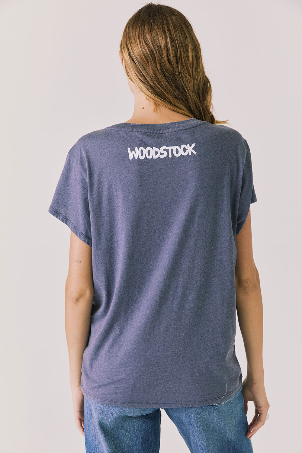 Back view of woman wearing Chaser’s slate blue Woodstock Love Revolution Tee with Woodstock in white text on a plain background (licensed music collaboration)