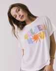 Front view of a woman wearing Chaser's white Golden Hour Tee with colorful graphic design on a plain background