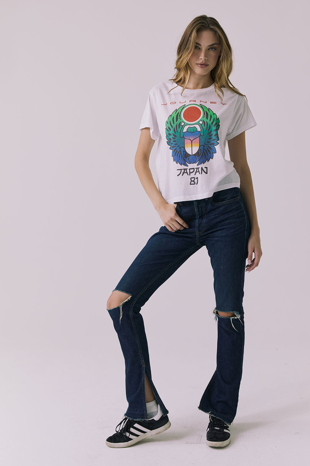 Full front view of a woman wearing Chaser's white Journey 1980 Tour Tee with a colorful graphic design and ripped denim on a plain background