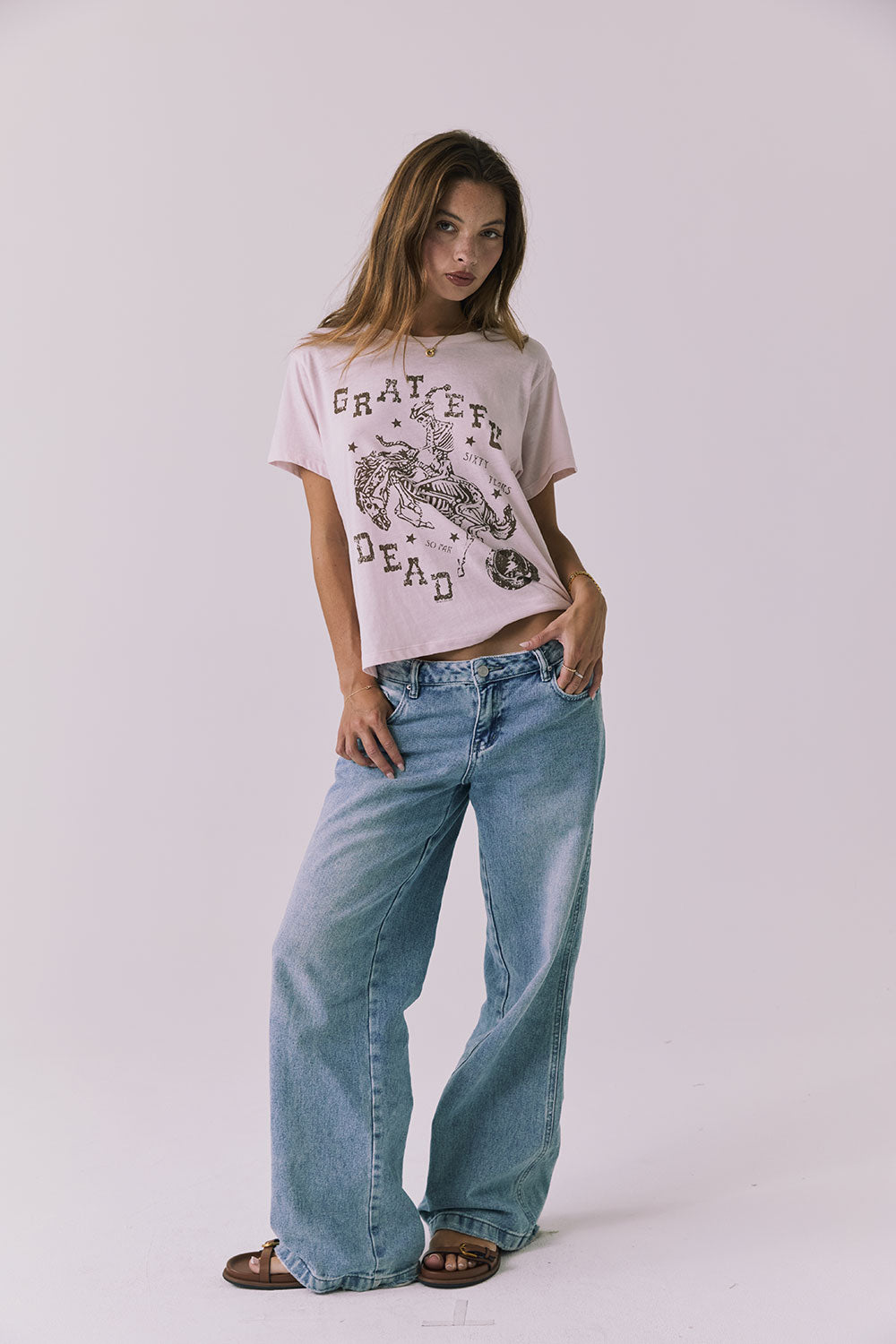 Full body front view of a woman wearing Chaser's the Grateful Dead 60 Years so Far pink tee with a graphic design and blue jeans on a plain background