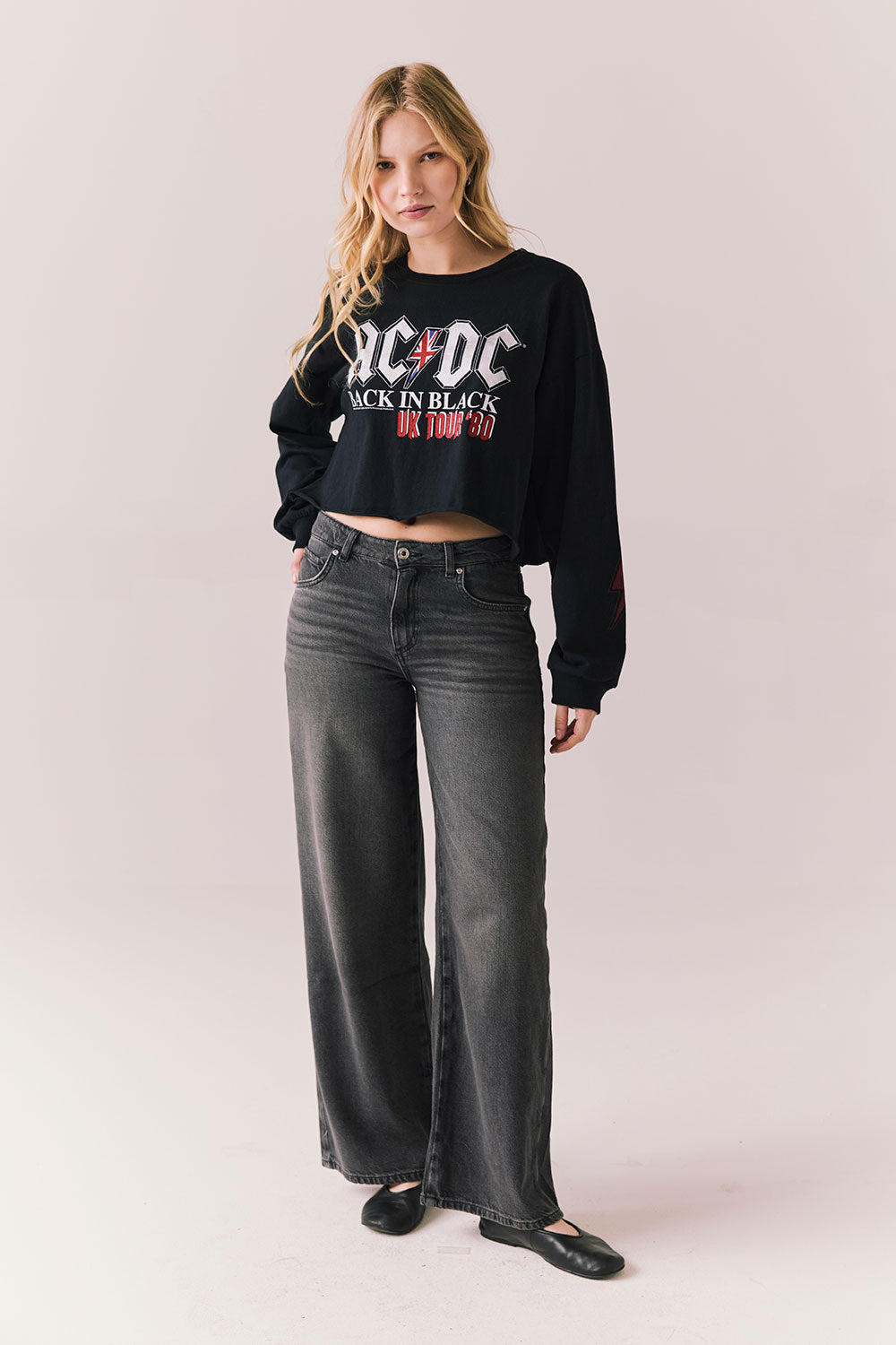 Full body view of model wearing AC/DC UK Tour ’80 Tee in True Black with cropped long-sleeve fit and vintage tour poster graphic paired with jeans on plain background