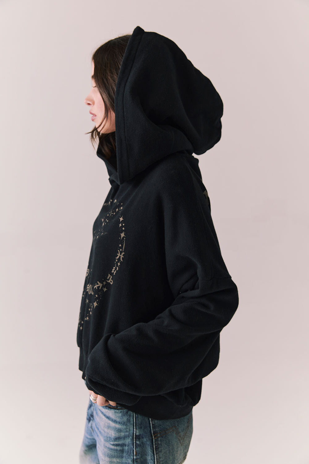 Side view of The Grateful Dead We Are Everywhere Hoodie in True Black with cropped boxy fit, hood up, and skull graphic made from stars and zodiac signs against a plain background