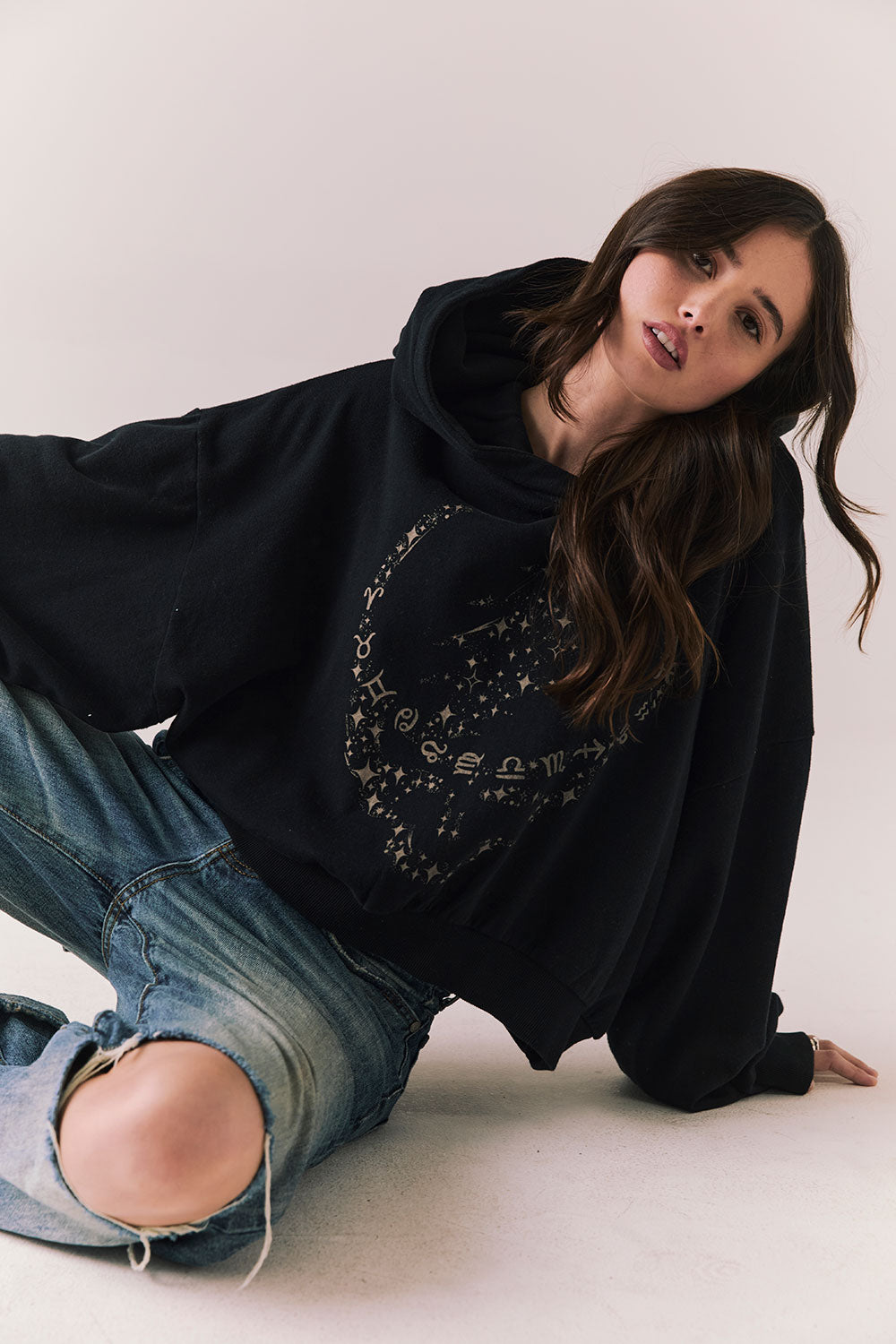 Front view of model wearing the The Grateful Dead We Are Everywhere Hoodie in True Black with cropped boxy fit and skull graphic made from stars and zodiac signs with denim sitting against a plain background