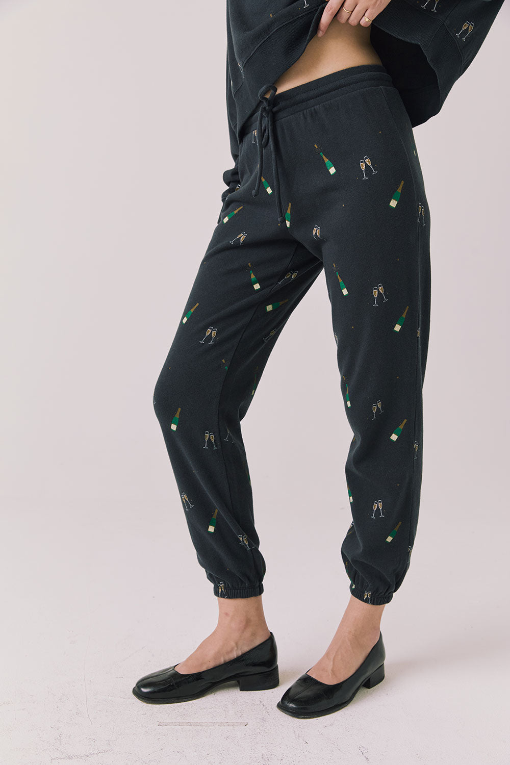 Side view of woman wearing Chaser’s black Champagne Party Joggers with allover champagne bottle and glass graphics on a light grey background