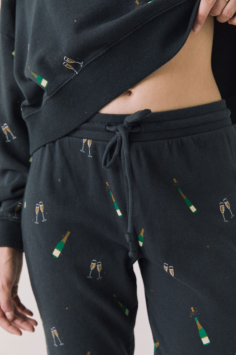 Front view of woman wearing Chaser’s black Champagne Party Joggers with allover champagne bottle and glass graphics on a light grey background