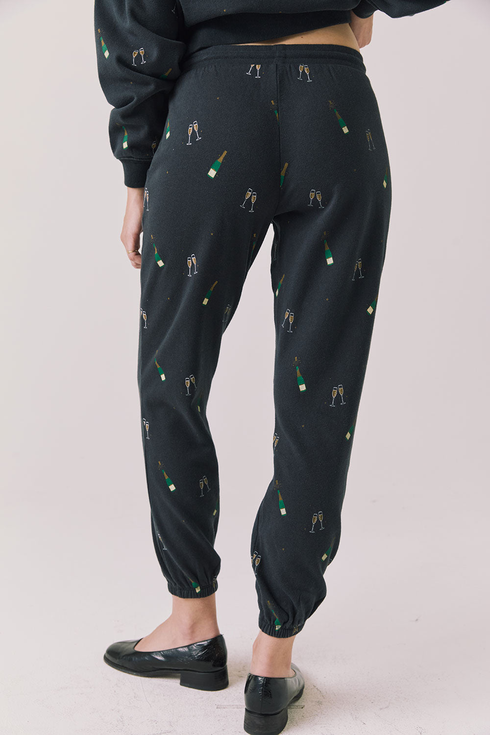 Back view of woman wearing Chaser’s black Champagne Party Joggers with allover champagne bottle and glass graphics on a light grey background