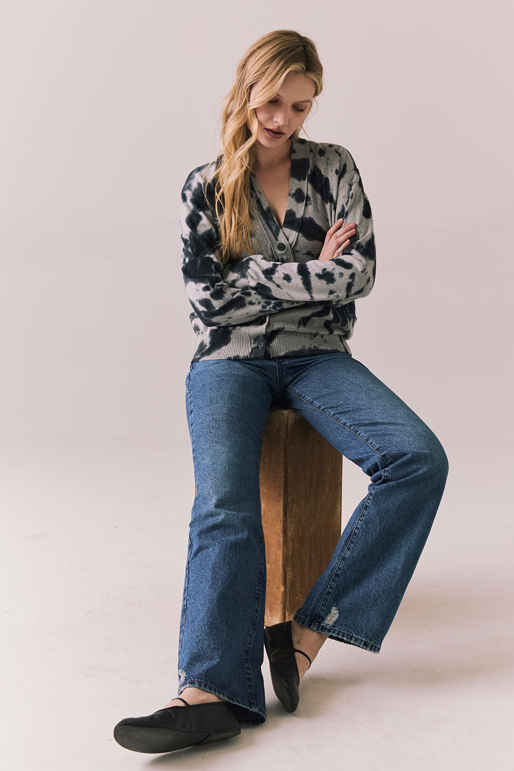 Full body view of Woman sitting on a wooden stool wearing the black tie dye hoodie and blue jeans against a plain background