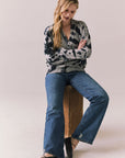 Full body view of Woman sitting on a wooden stool wearing the black tie dye hoodie and blue jeans against a plain background