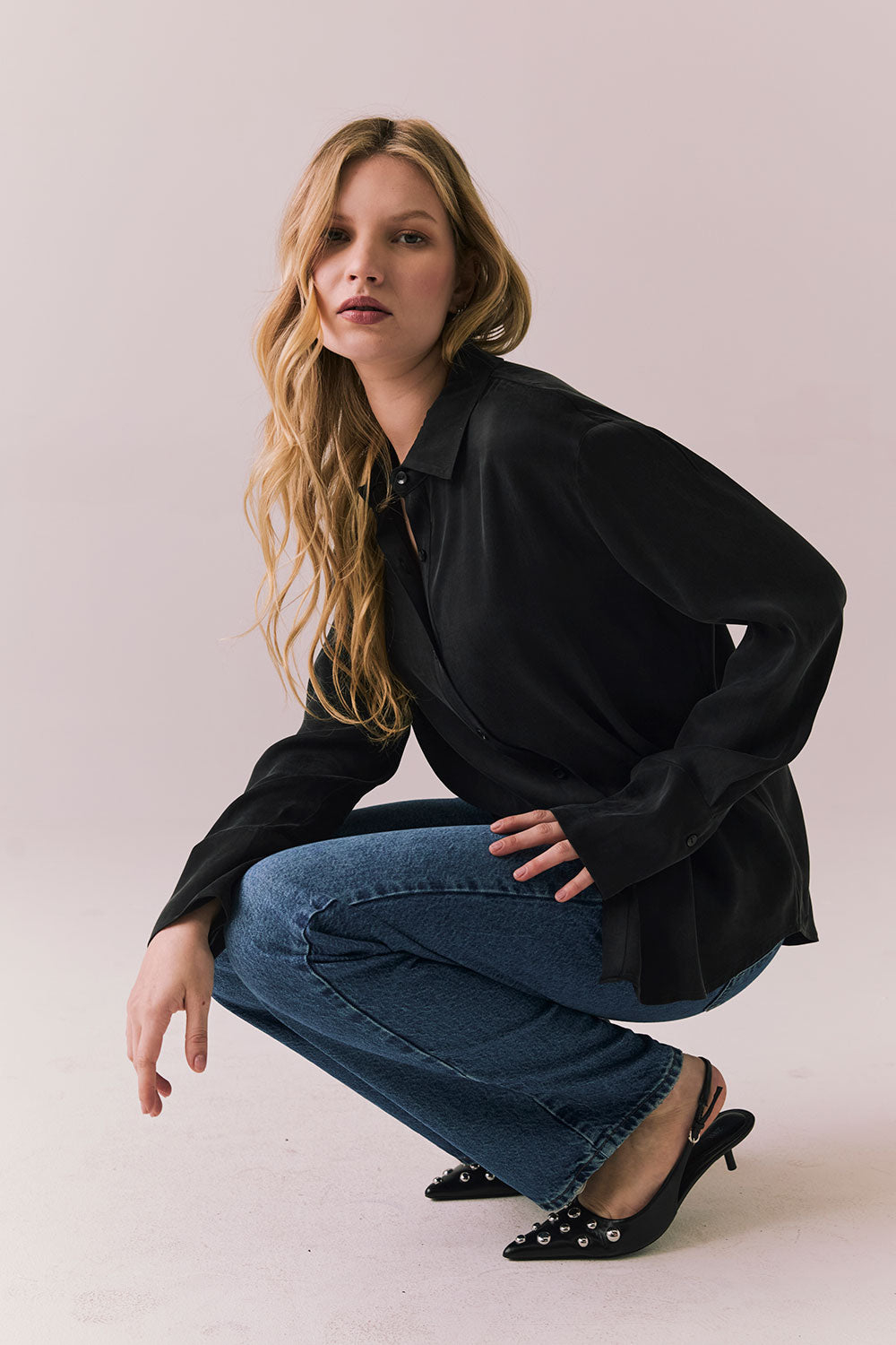 Full side view of a model wearing the Helenaa button-down shirt in True Black with collared neckline and oversized silhouette, styled with relaxed jeans and crouched against a plain backdrop.