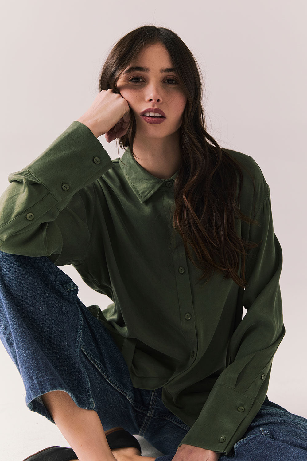 Front view of a model wearing the Helenaa button-down shirt in bronze green with collared neckline and relaxed silhouette, styled with blue jeans, sitting on the floor against a plain background.