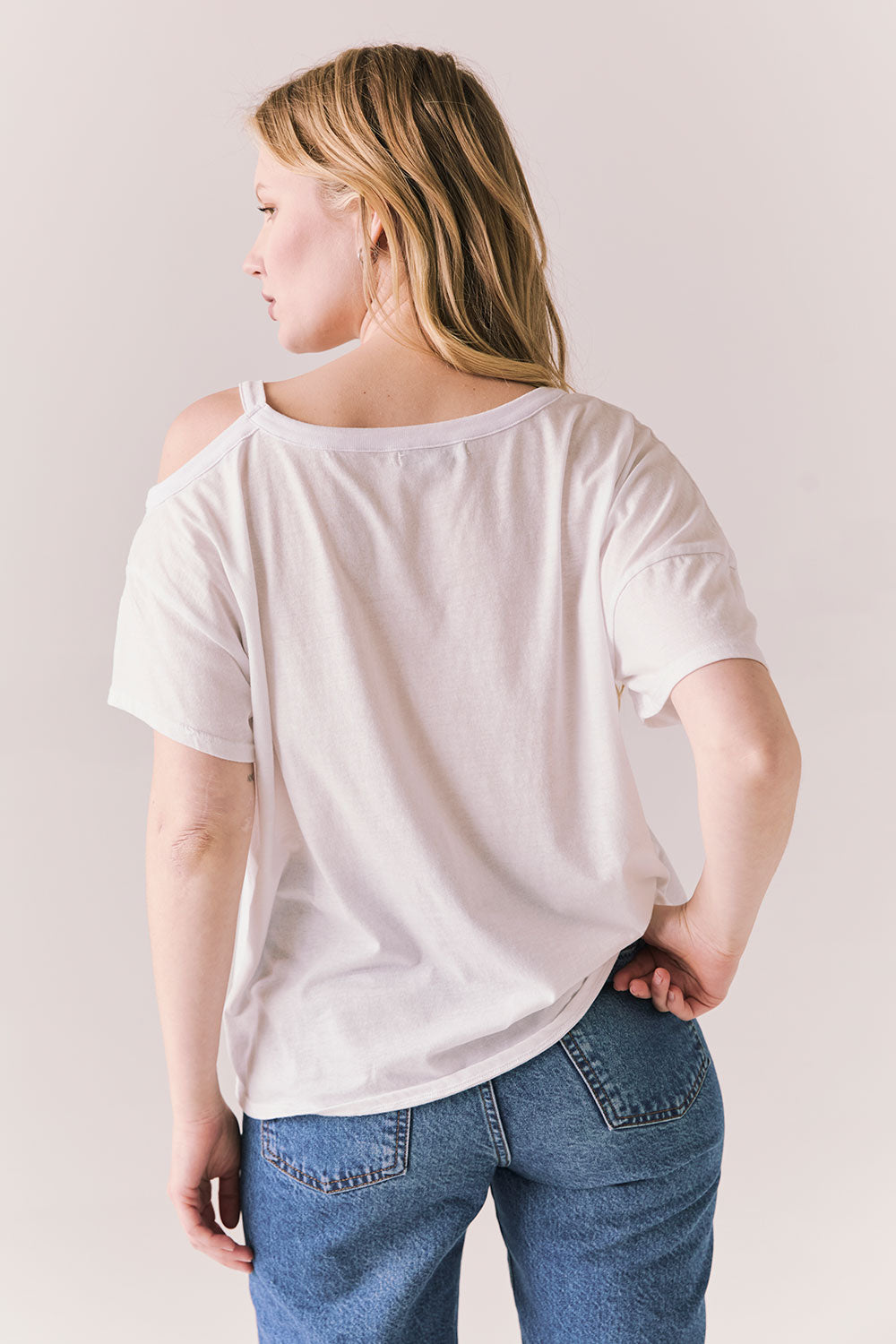 Back view of Northridge Tee in white with cold shoulder cutout and flowy fit and blue jeans against a plain background