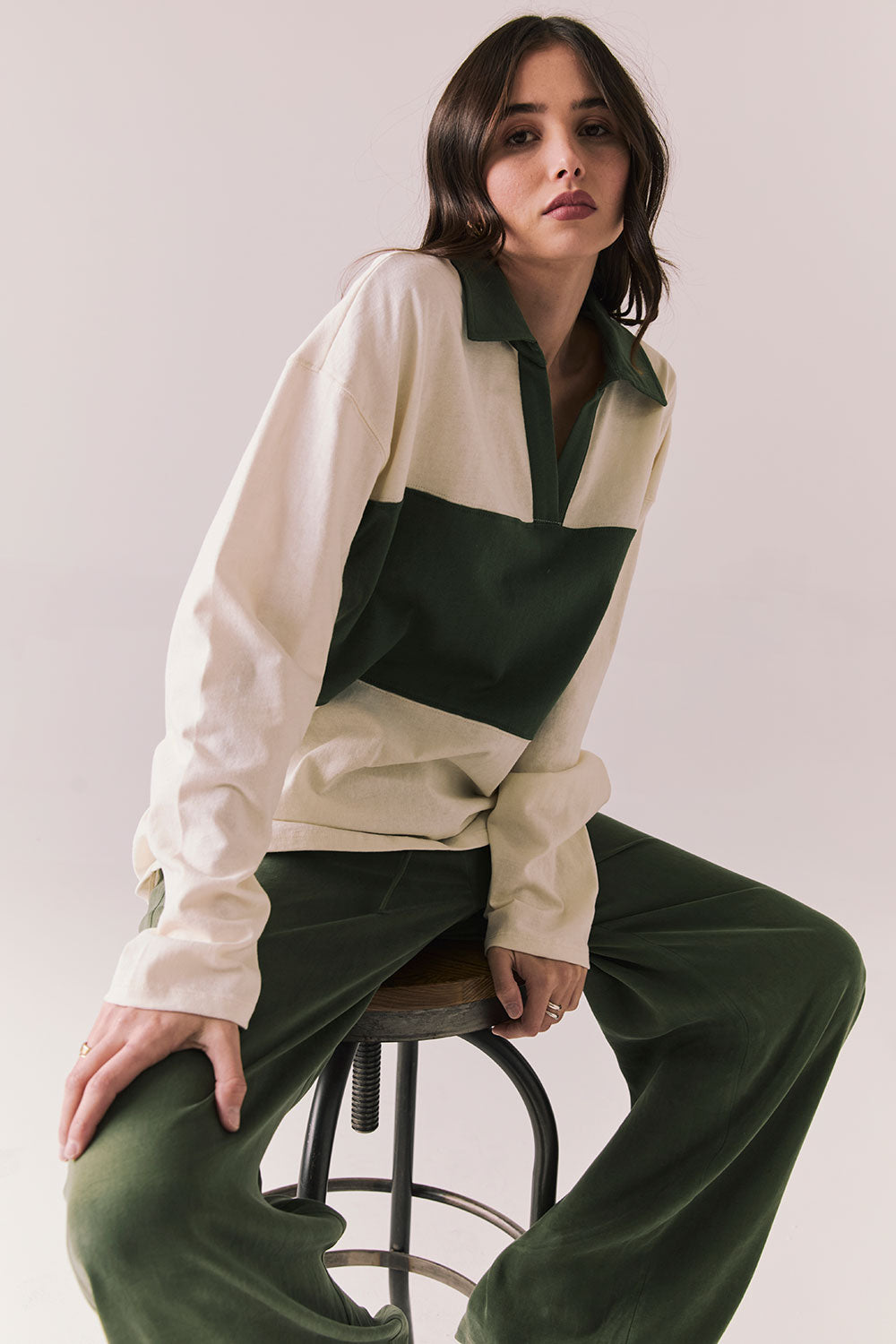 Woman wearing the color-blocked Chico Long Sleeve White and Green Rib Pullover and green pants sitting on a stool against a plain background