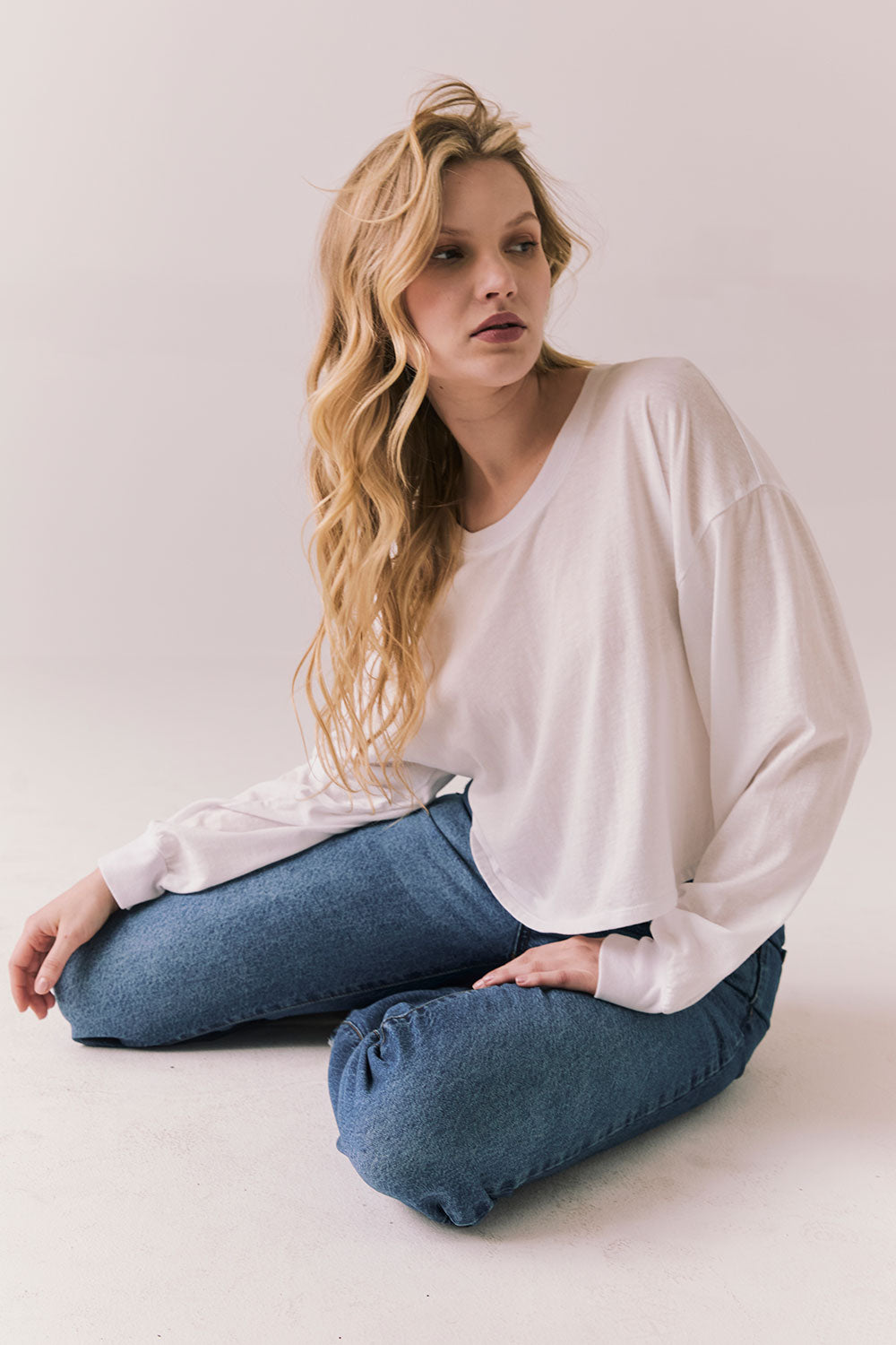 Woman sitting on the floor wearing the Hartt long sleeve in white with cotton texture and clean silhouette and blue jeans against a plain background