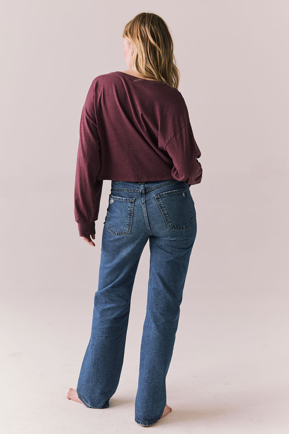 Back full body view of model wearing the Hartt long sleeve tee in fig (deep purple) with clean silhouette and blue jeans, against a plain background.