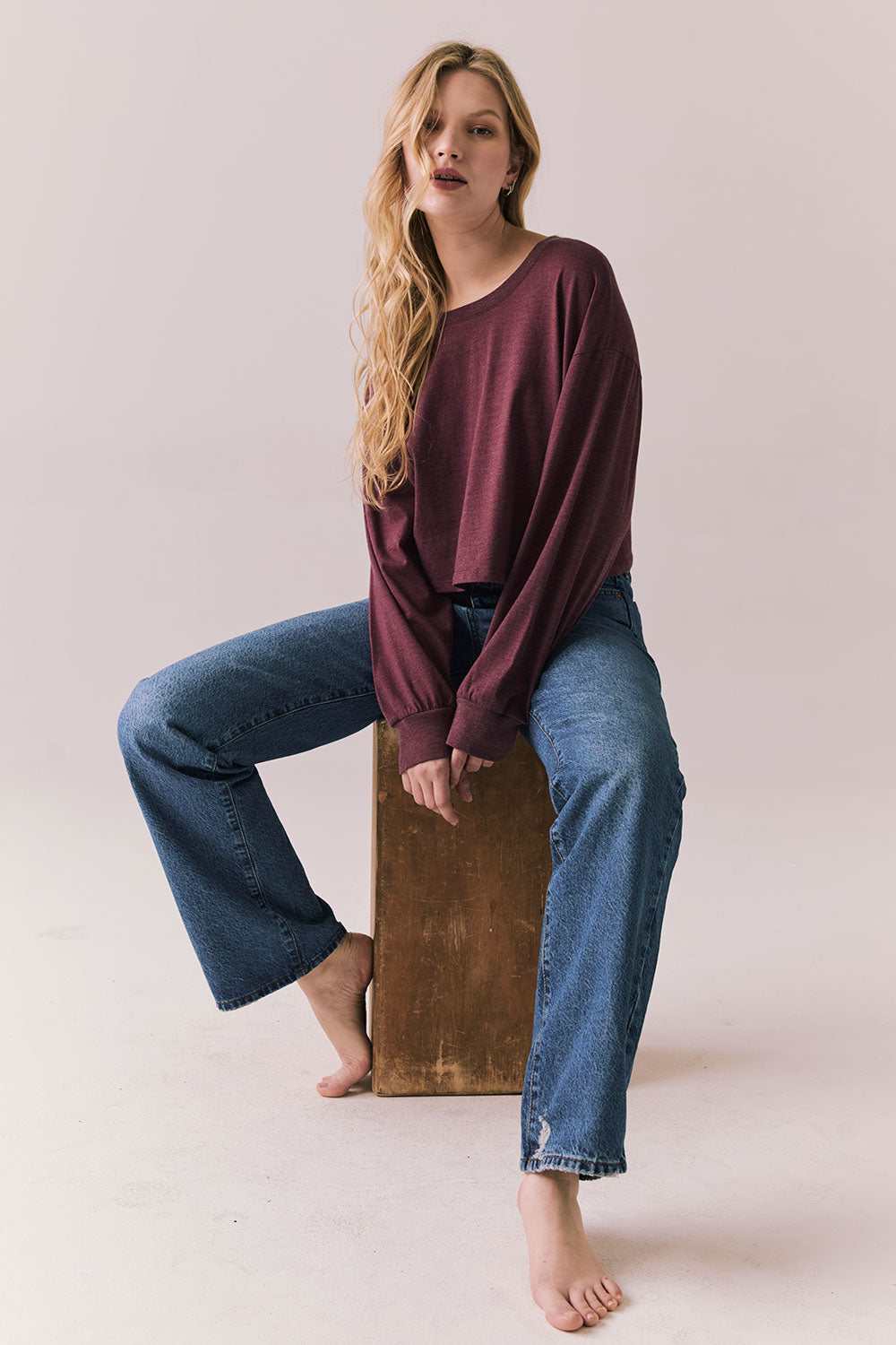 Full view of model wearing the Hartt long sleeve tee in fig (deep purple) with clean silhouette and blue jeans, seated on a wooden block against a plain background.