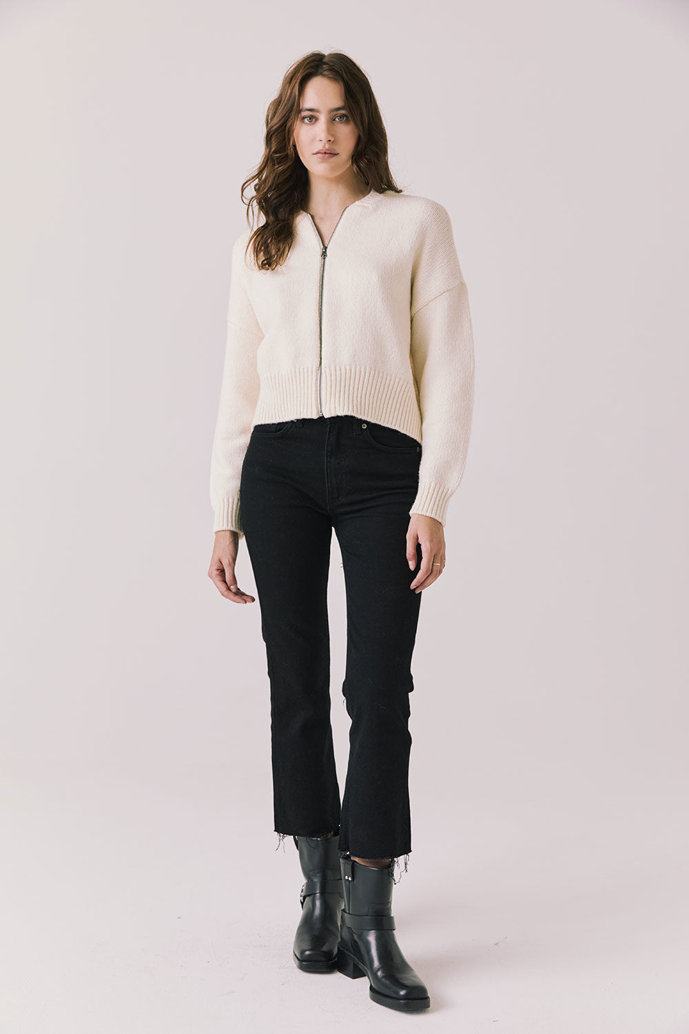 Full body view of woman wearing Chaser’s Warm White Luck Zip Up Cardigan Sweater with black pants and black boots on a white background