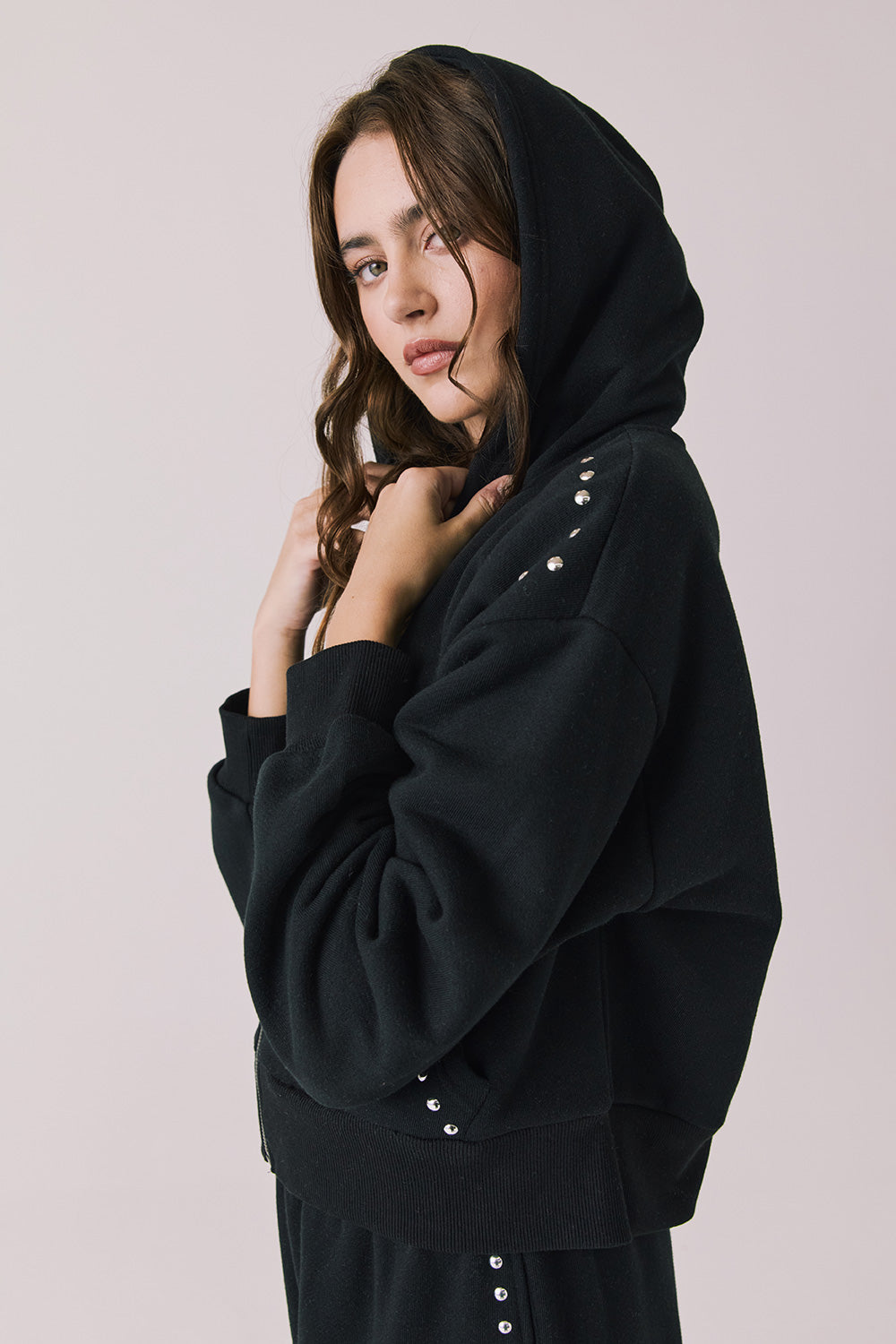 Side view of woman wearing Chaser’s Kiki True Black Zip Up Hoodie with sequin embellishments, hood up, and matching Daze True Black Joggers on a white background