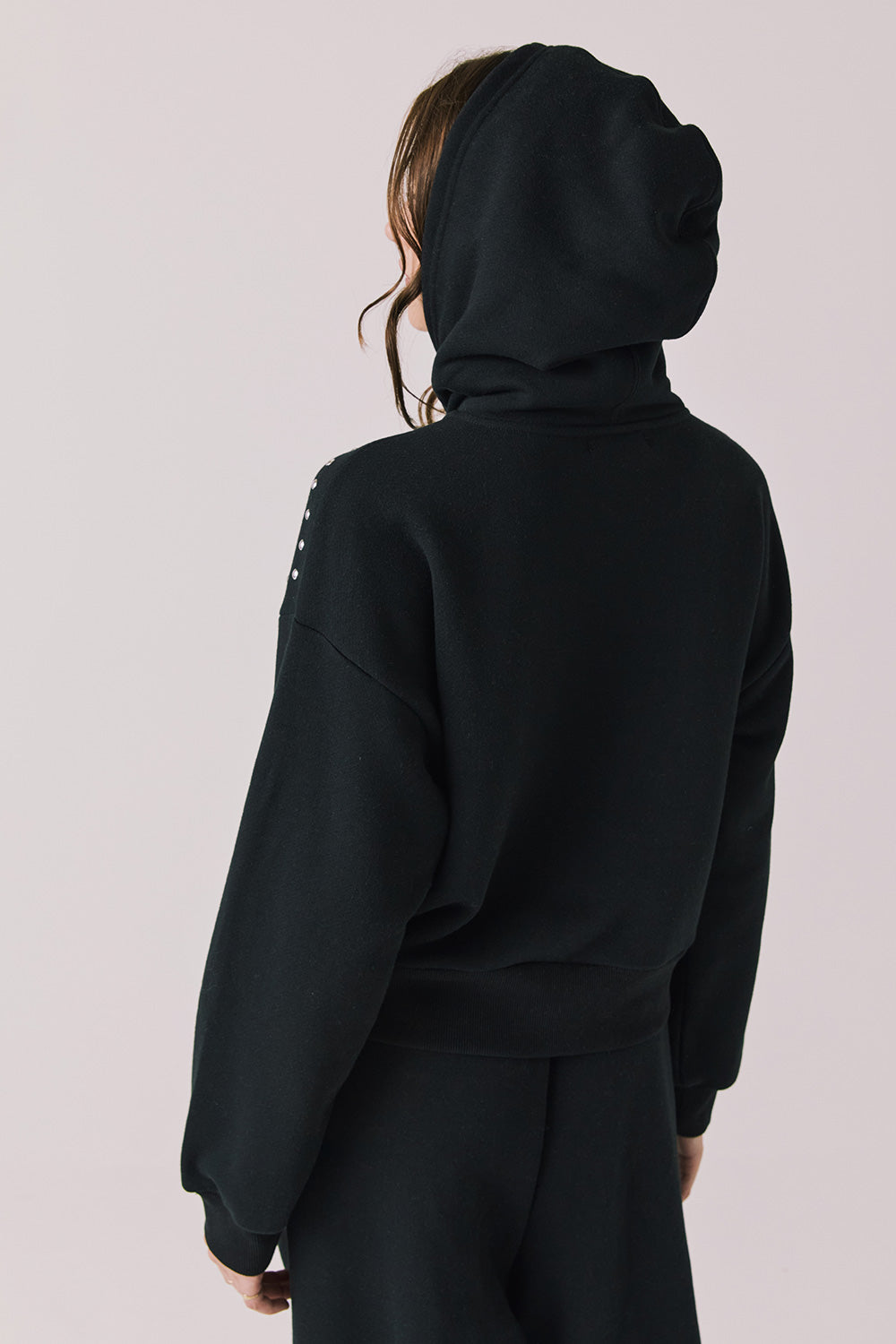 Back view of woman wearing Chaser’s Kiki True Black Zip Up Hoodie with sequin accents with hood up on the shoulders and pockets on a white background