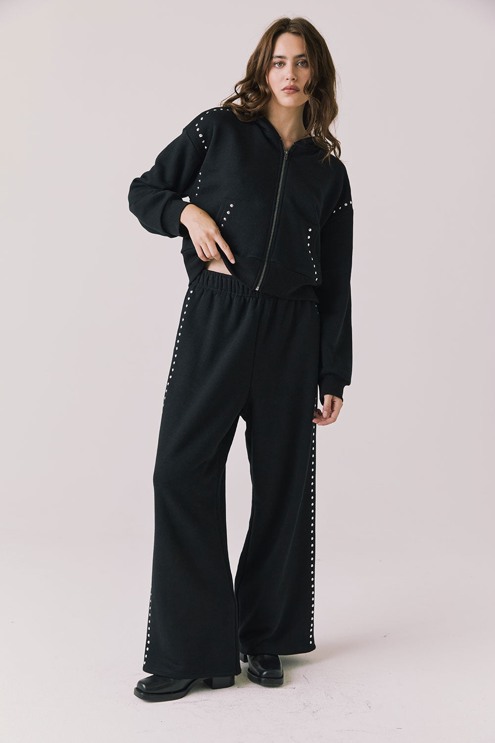 Full body view of woman wearing Chaser’s Daze True Black Joggers and matching Kiki True Black Zip Up Hoodie with sequin details on a white background