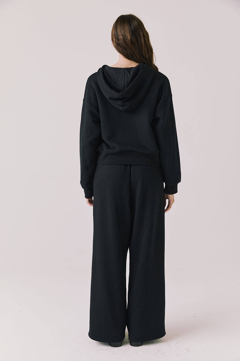 Full body back view of woman wearing Chaser’s Daze True Black Joggers and matching Kiki True Black Zip Up Hoodie with sequin details on a white background
