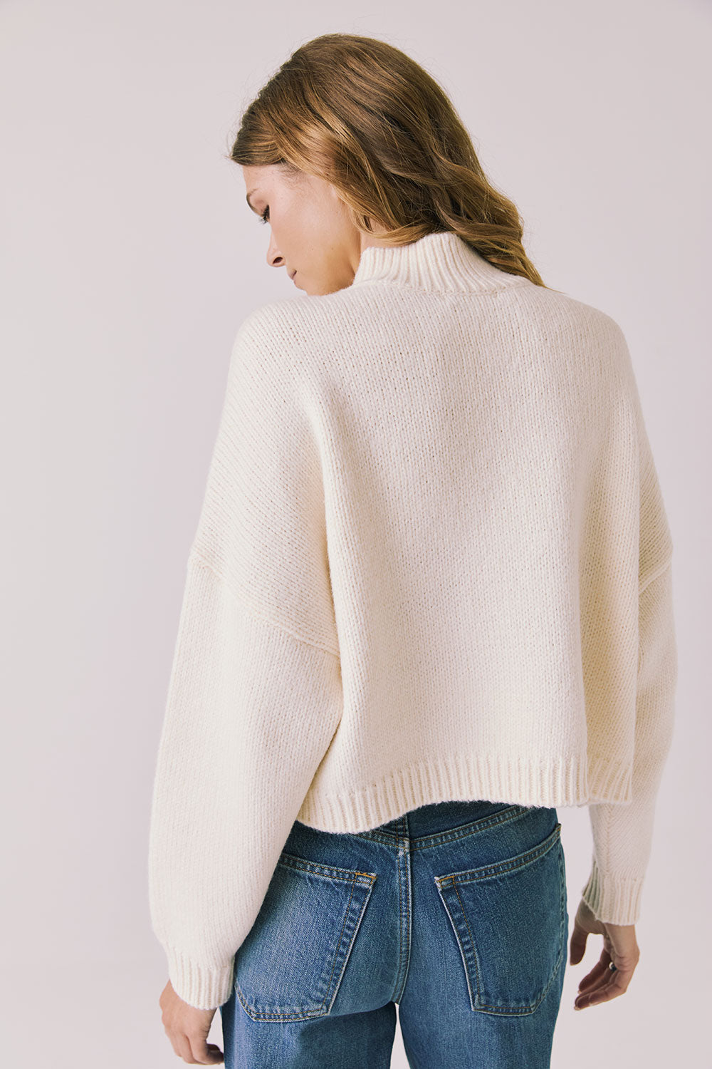 Back view of woman wearing Chaser’s Frankie Warm White Pullover Sweater with blue jeans on a white background