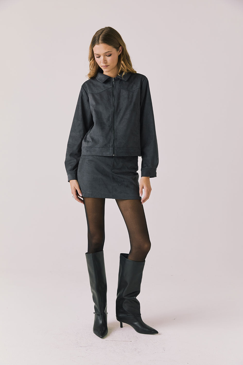 Full body view of woman wearing Chaser’s Dusk True Black Jacket with matching Brooke True Black Mini Skirt and black knee-high boots on a light background