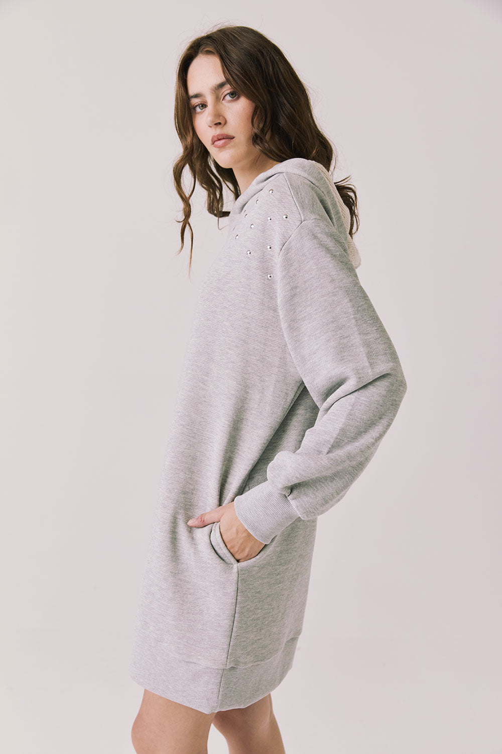Side view of woman wearing Chaser's Aster Heather Grey Hoodie Dress with hand in pocket on a plain background