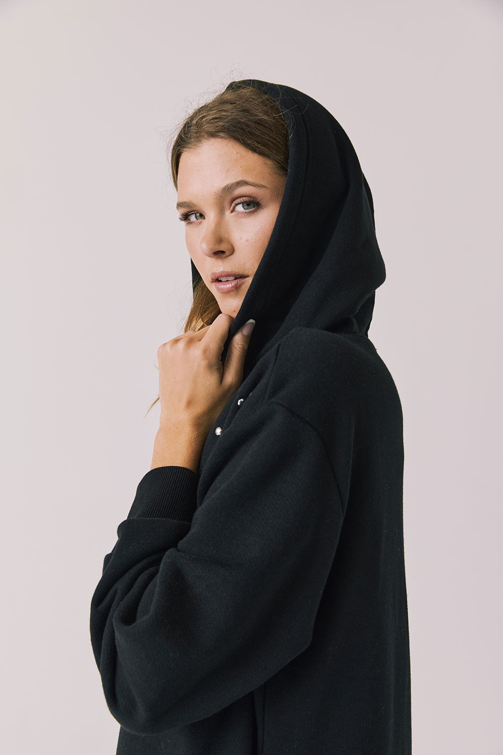 Side view of woman wearing Chaser Aster True Black Hoodie Dress with decorative elements on shoulders with hoodie up on a plain background