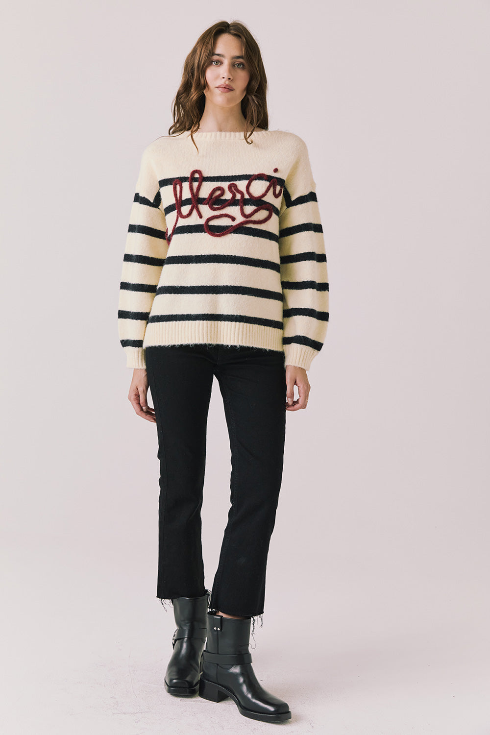 Full body view of woman wearing Chaser’s striped Merci Long Sleeve Sweater with “merci” burgundy red text, black pants, and black boots on a white background