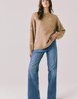 Full body view of woman wearing Chaser’s Love Me Long Sleeve Beige Sweater and blue jeans on a plain background