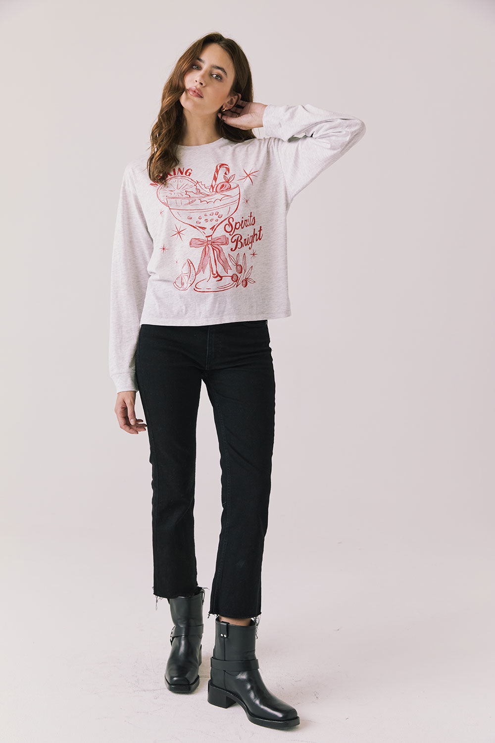 Full body view of woman wearing Chaser’s Spirits Bright Light Grey Long Sleeve with red graphics and text, black pants, and black boots on a light gray background