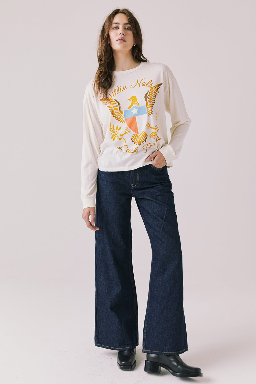 Full body view of woman wearing Chaser’s starry white Willie Nelson Luck Texas Long Sleeve with graphic design and dark blue jeans on a light background (licensed band collaboration)