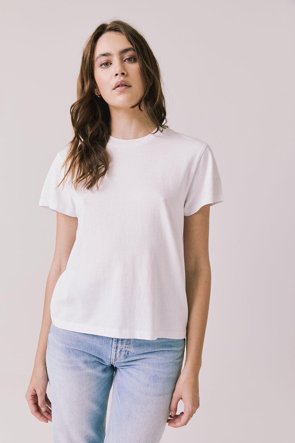 Front view of woman wearing Chaser’s Cosmo White Tee with short sleeves and light blue jeans on a plain background