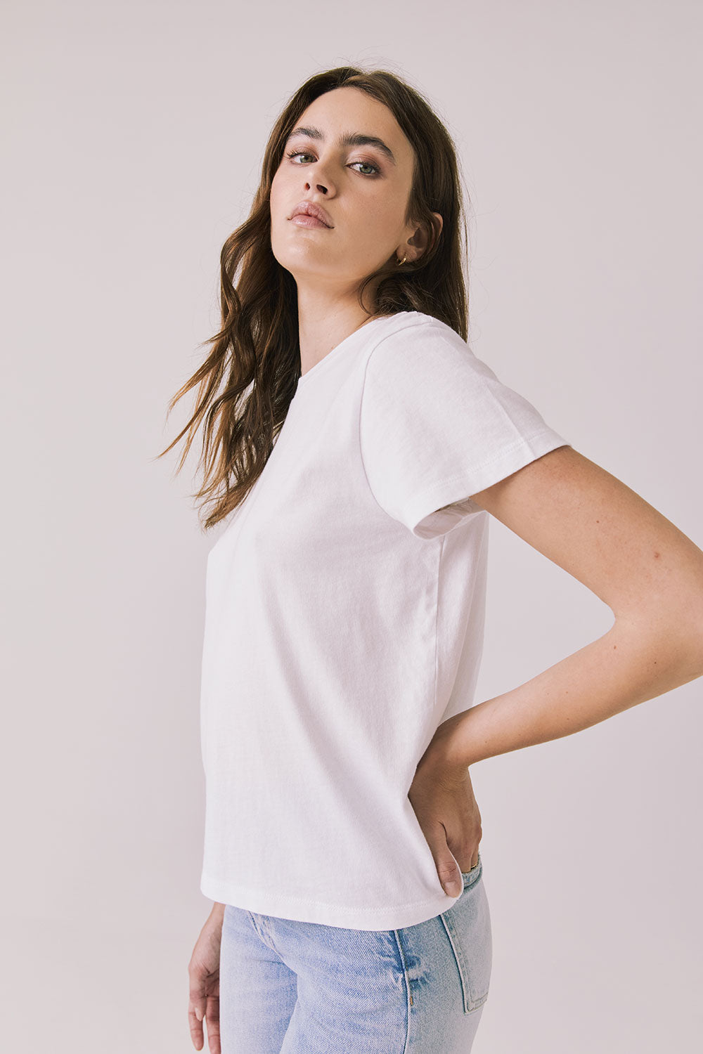 Side view of woman wearing Chaser’s Cosmo White Tee with classic crew neckline and light blue jeans on a plain background