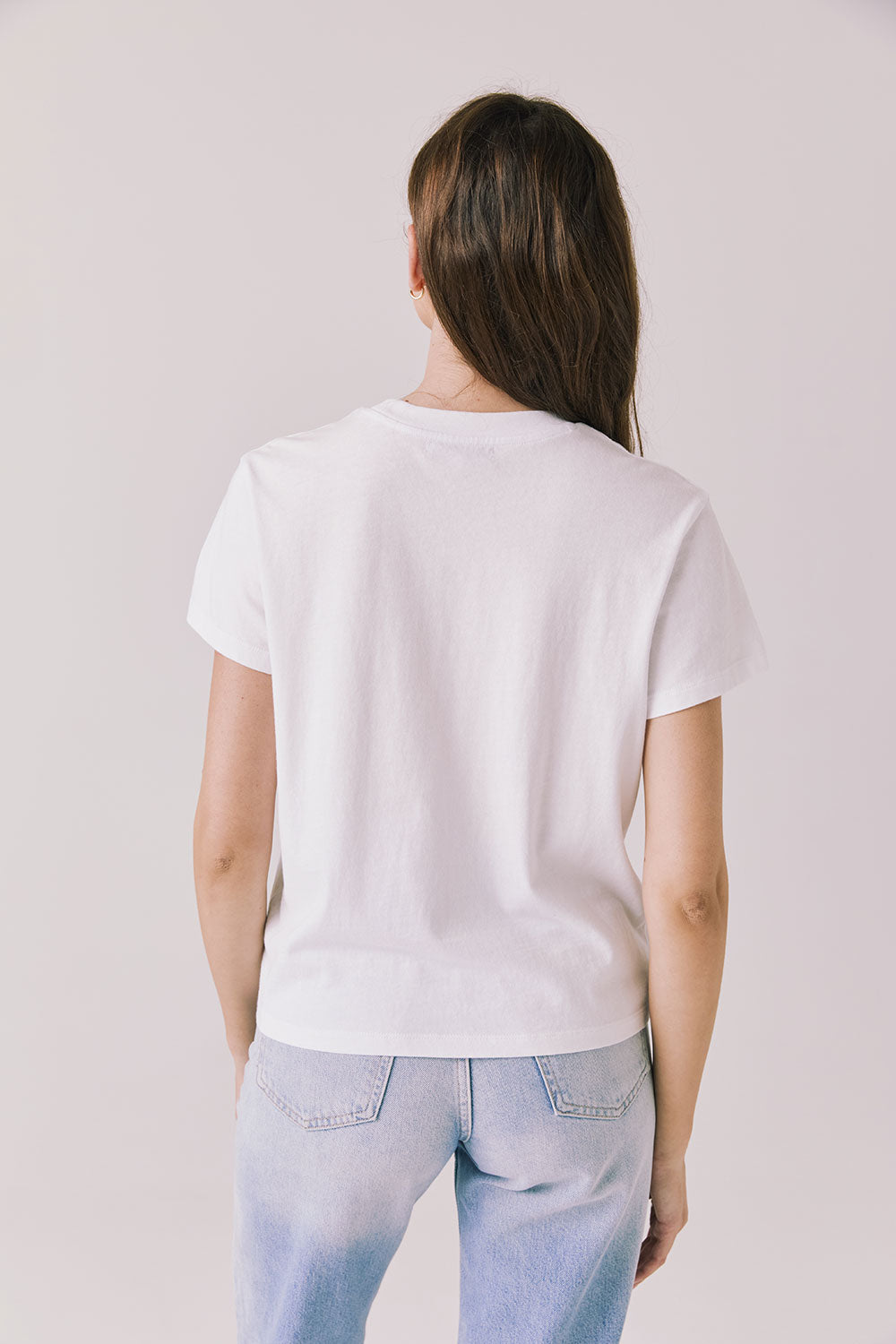 Back view of woman wearing Chaser’s Cosmo White Tee with relaxed fit and light blue jeans on a plain background