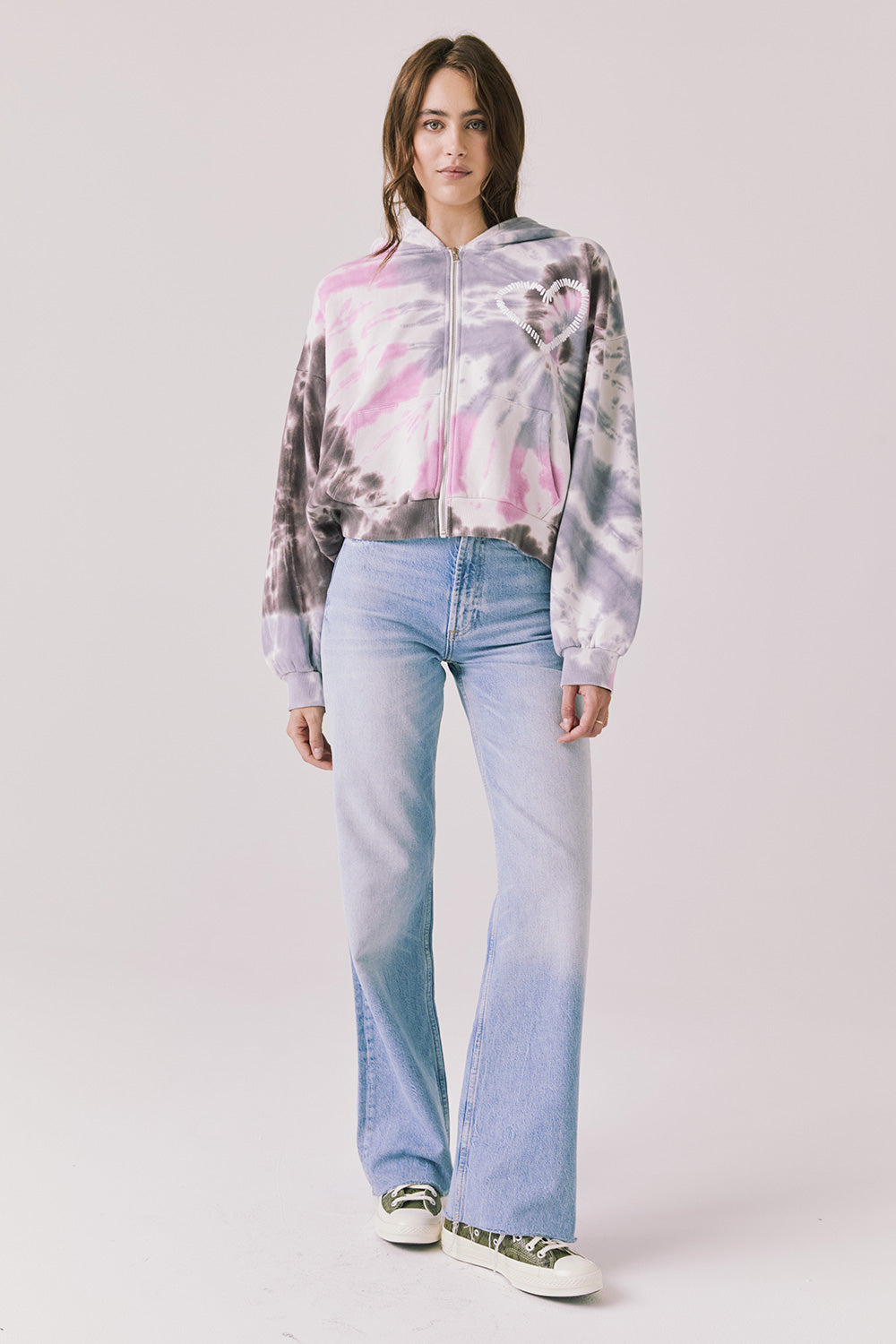 Full body view of woman wearing Chaser’s Heart Tie Dye Zip Up Hoodie with light blue jeans on a white background